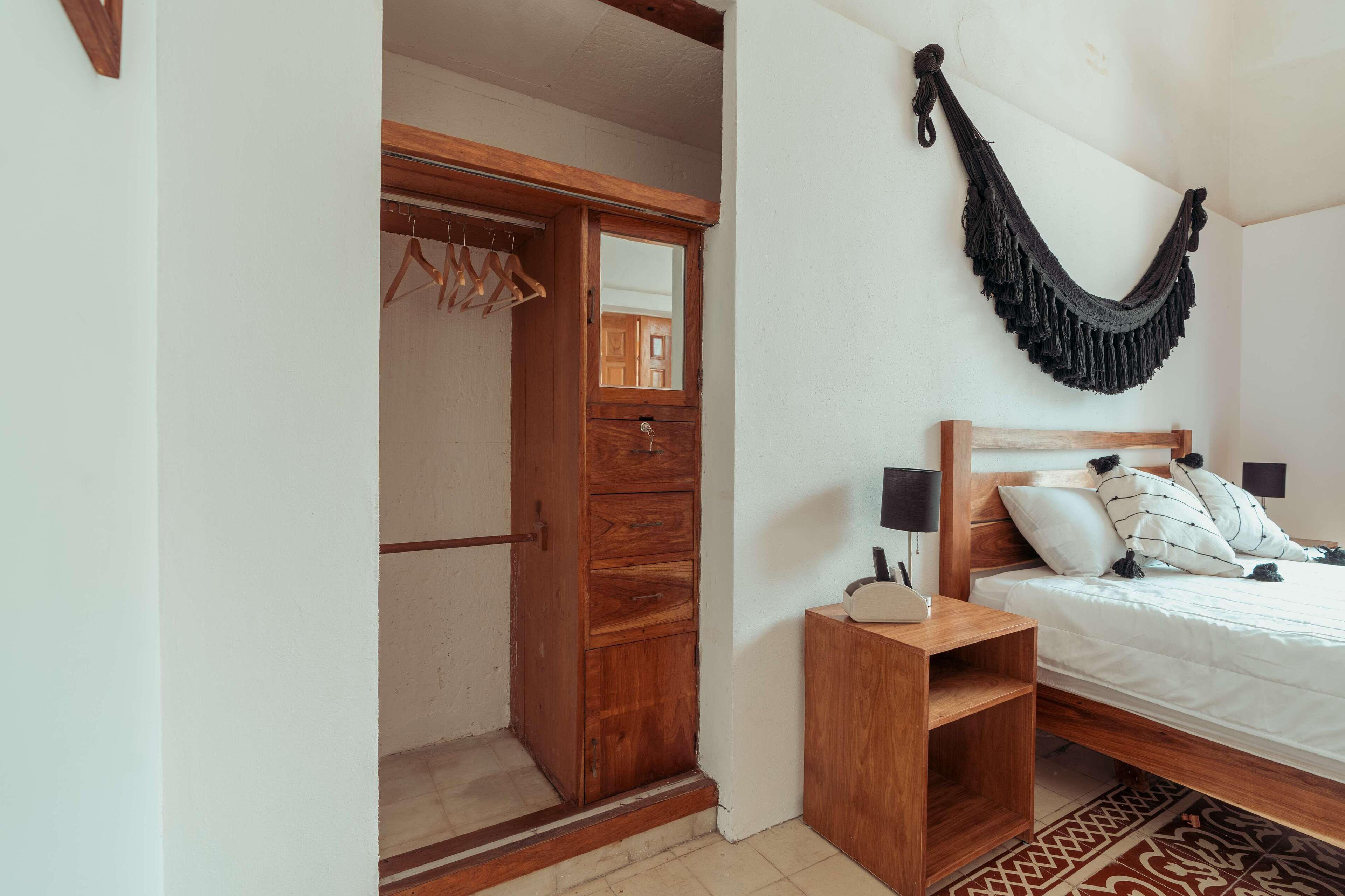 A bedroom with a wooden wardrobe, a bed with white bedding, a hanging black fringe ornament, and a bedside table with a lamp.