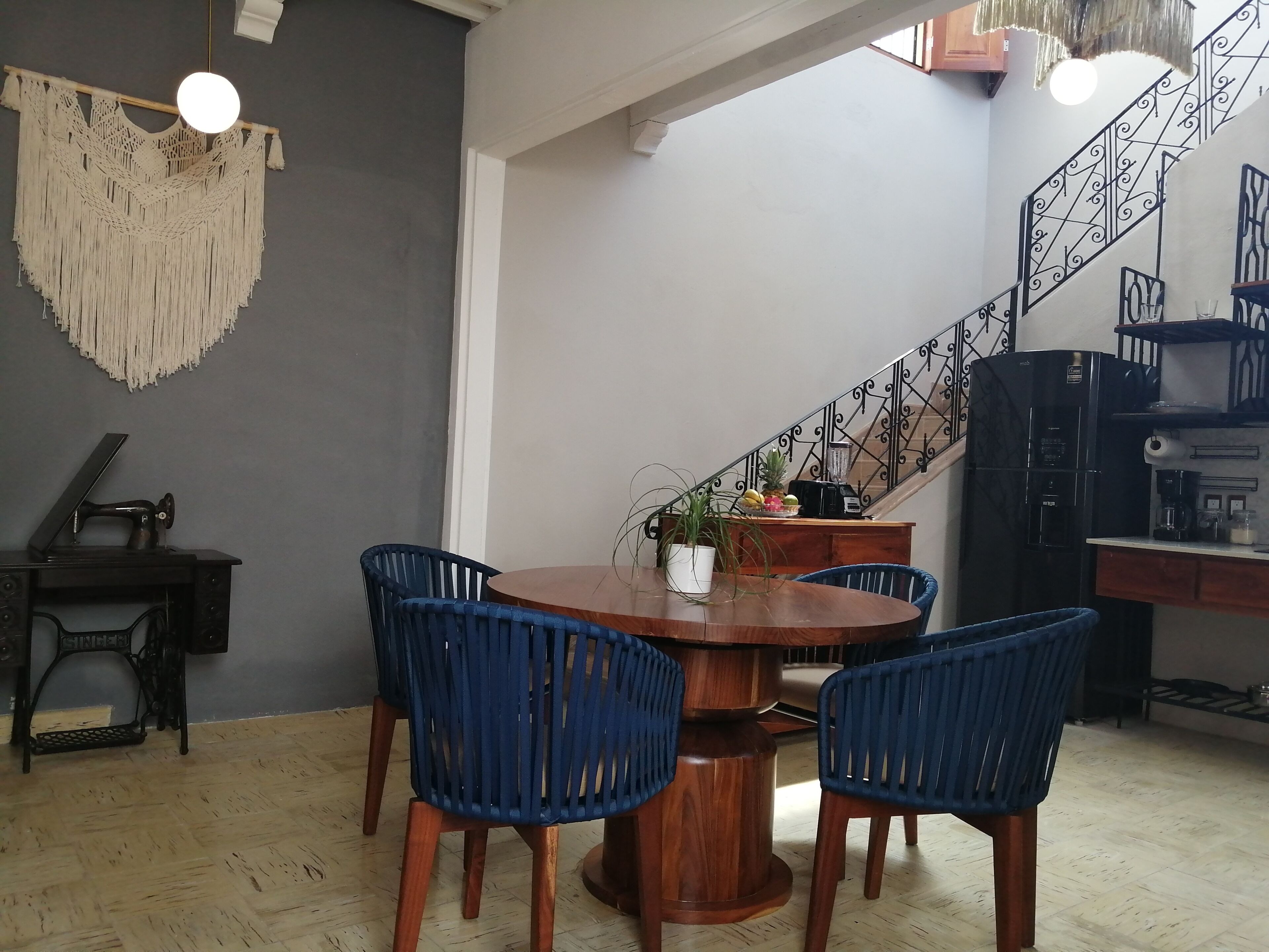 A dining area with a round wooden table, blue chairs, a sewing machine, and a macramé wall hanging.