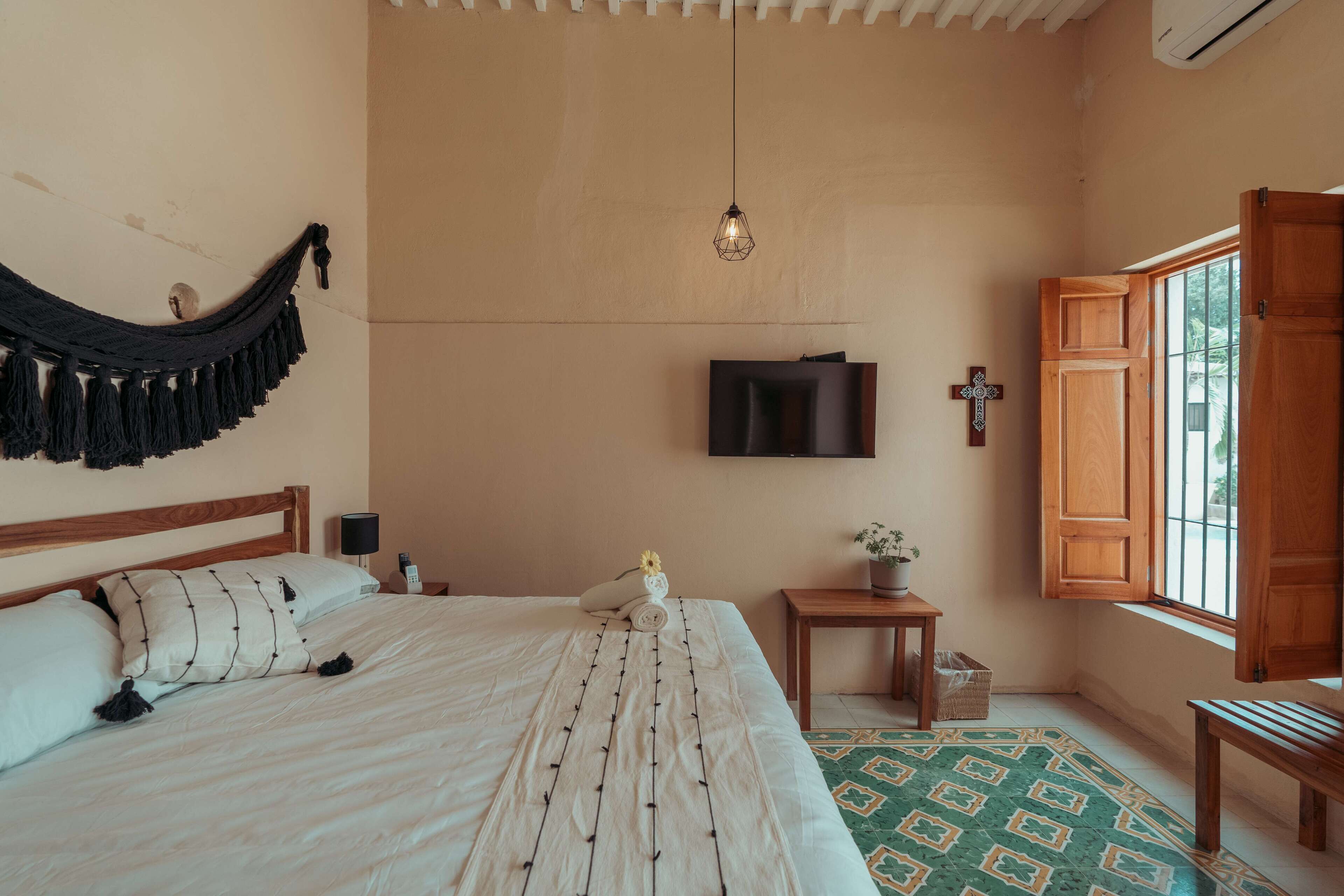 Un dormitorio con cama, hamaca, televisor, una cruz y ventana con persianas de madera.