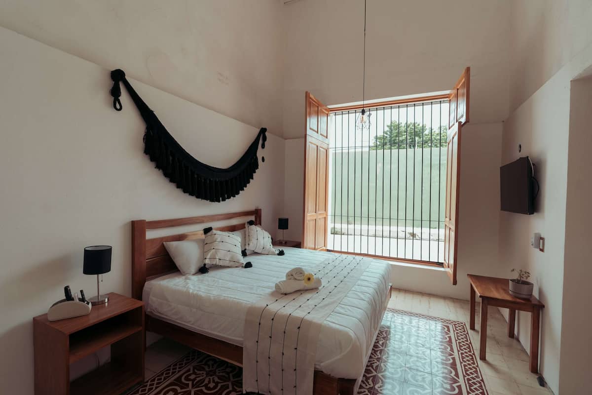 A bedroom with a bed, bedside table, lamp, and a window with a view of greenery.