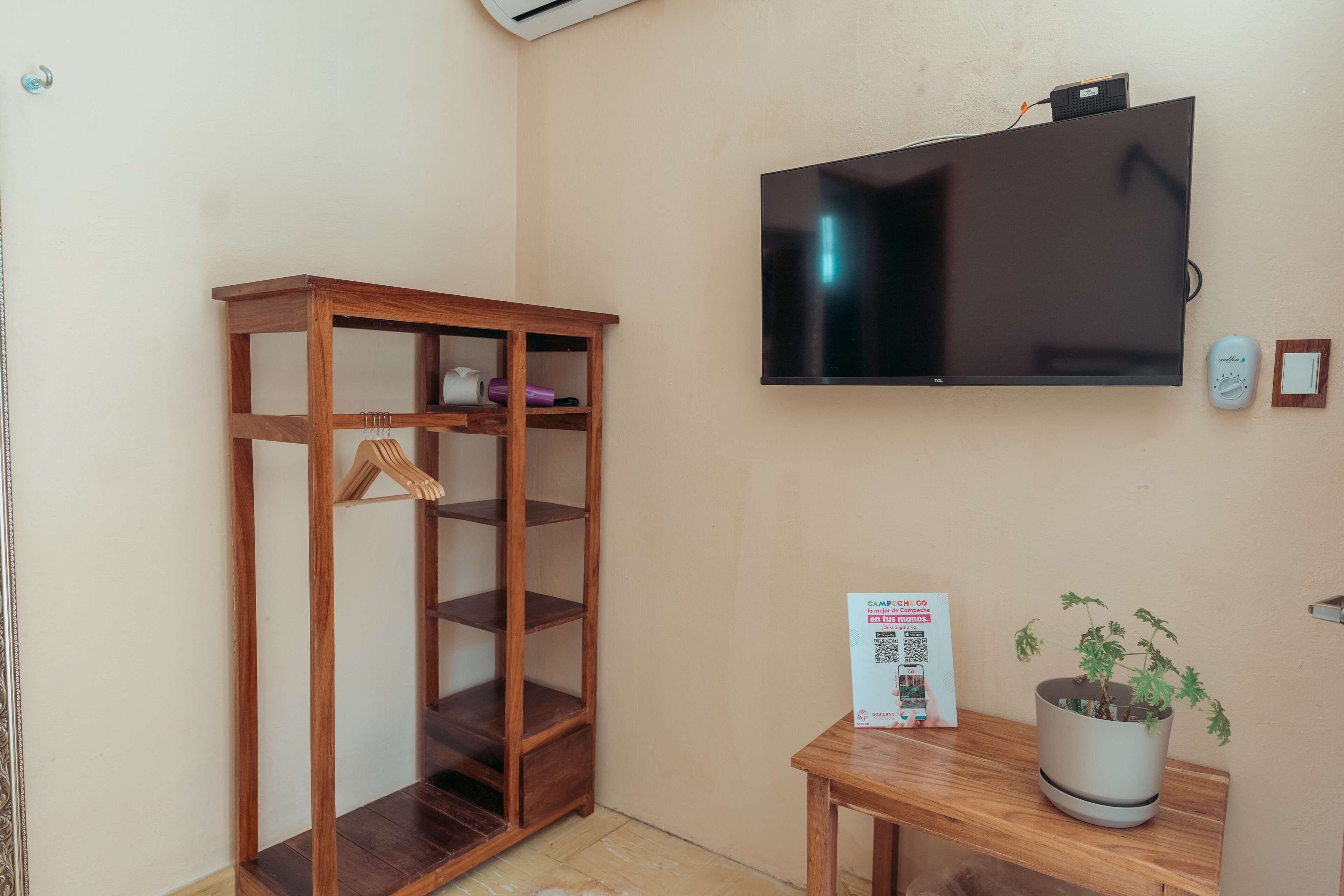 Une pièce comprenant un meuble en bois, un téléviseur fixé au mur, une petite table avec un pot de plante et un panneau avec un code QR.