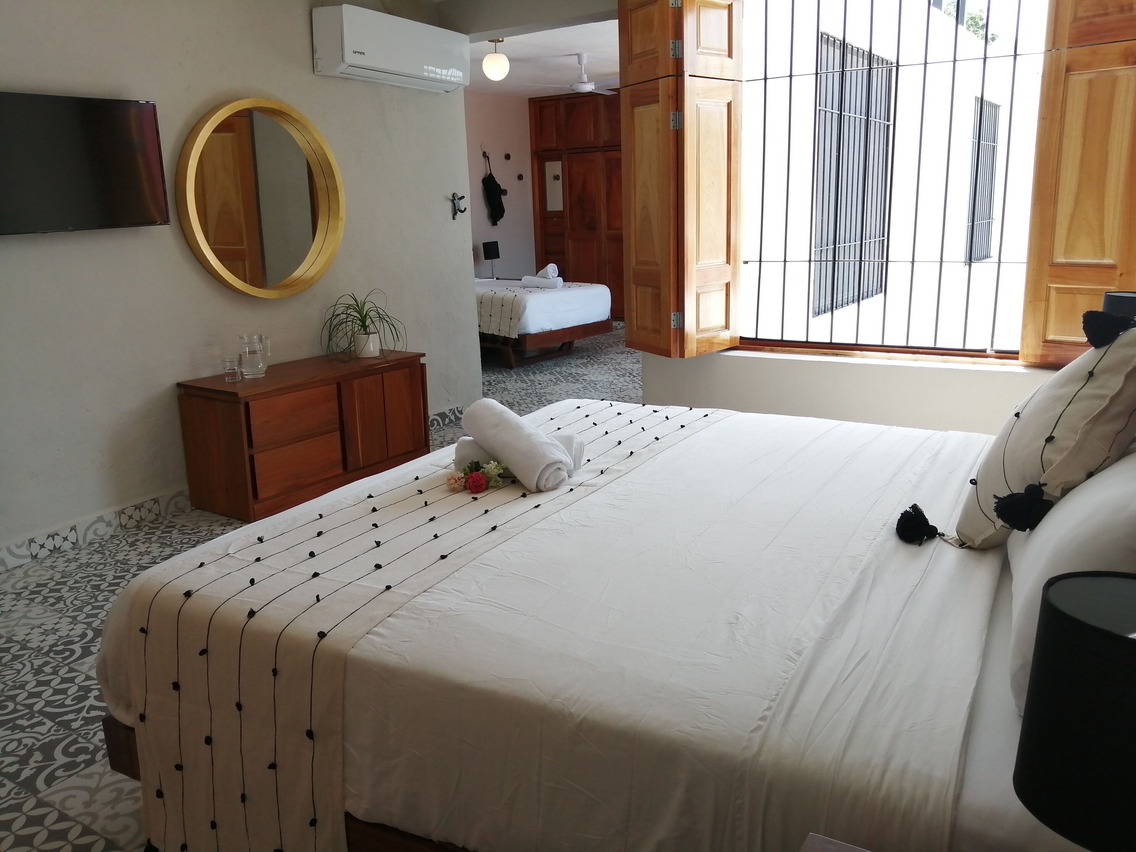 Un dormitorio con cama, cómoda de madera, espejo, equipo de aire acondicionado y ventana con barrotes.