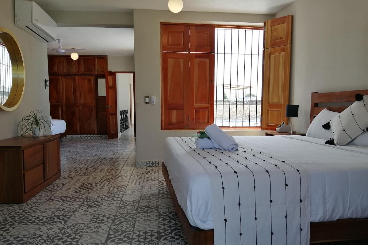 A hotel room with a bed, a wooden wardrobe, a bedside table with a lamp, a potted plant, and a view through a large window.