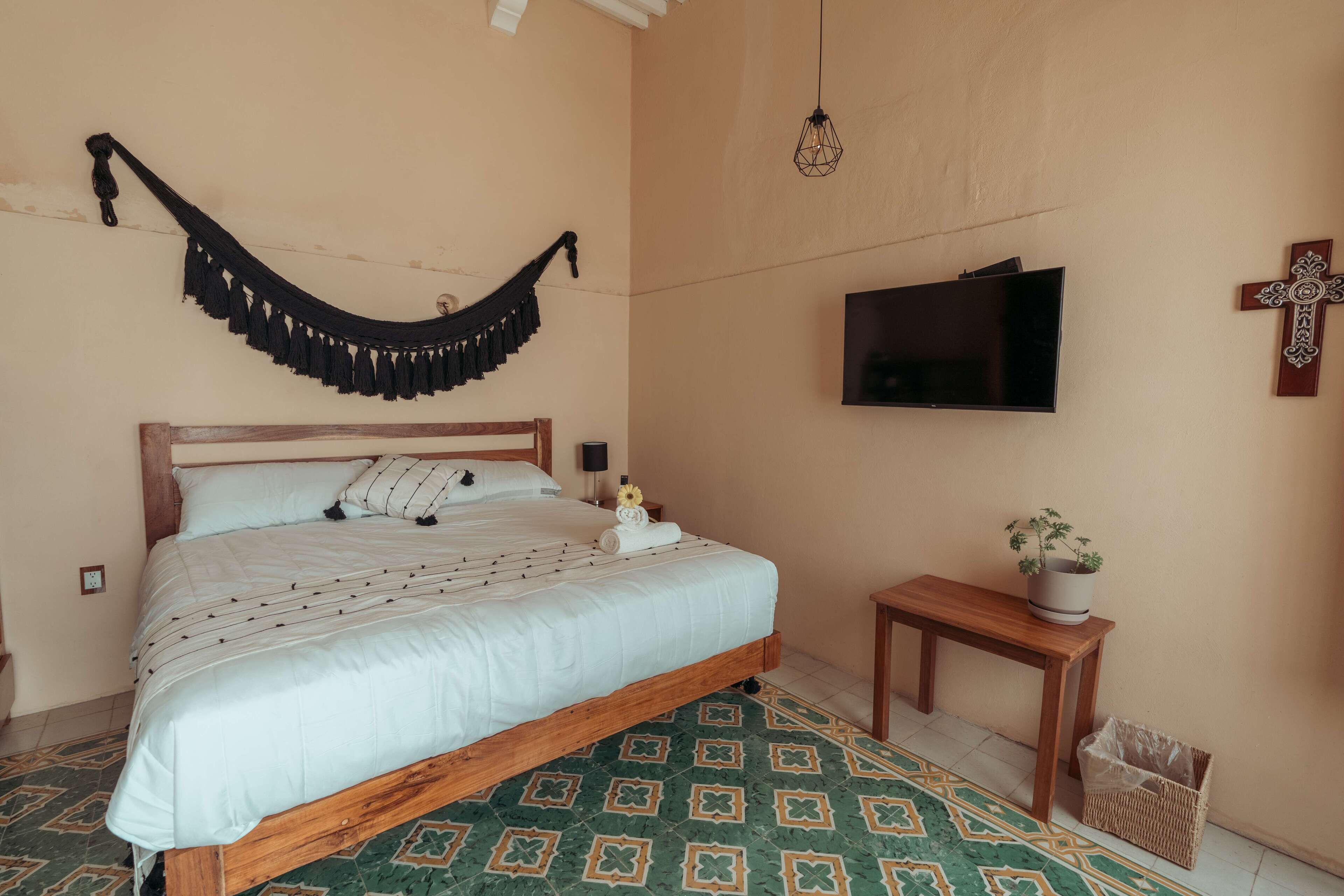 A neatly made bed with a wooden frame, a hanging black hammock, a small bedside table with a plant, and a television mounted on the wall.
