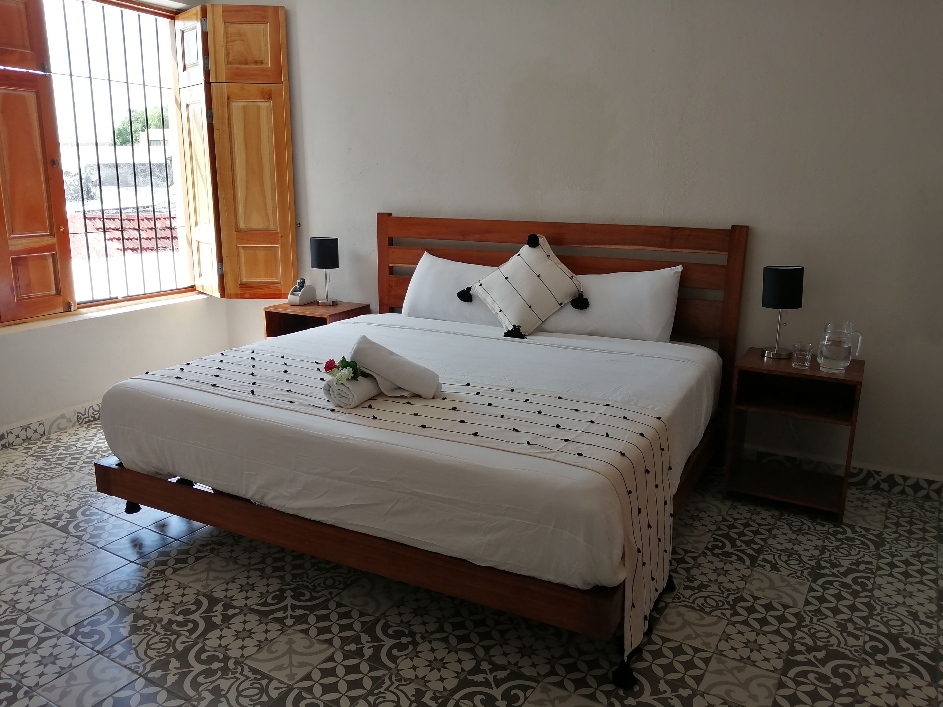 A neatly made bed with a white bedspread, a rolled towel, and a decorative pillow. There are two bedside tables with lamps and a water pitcher. The room has patterned floor tiles and a wooden door.