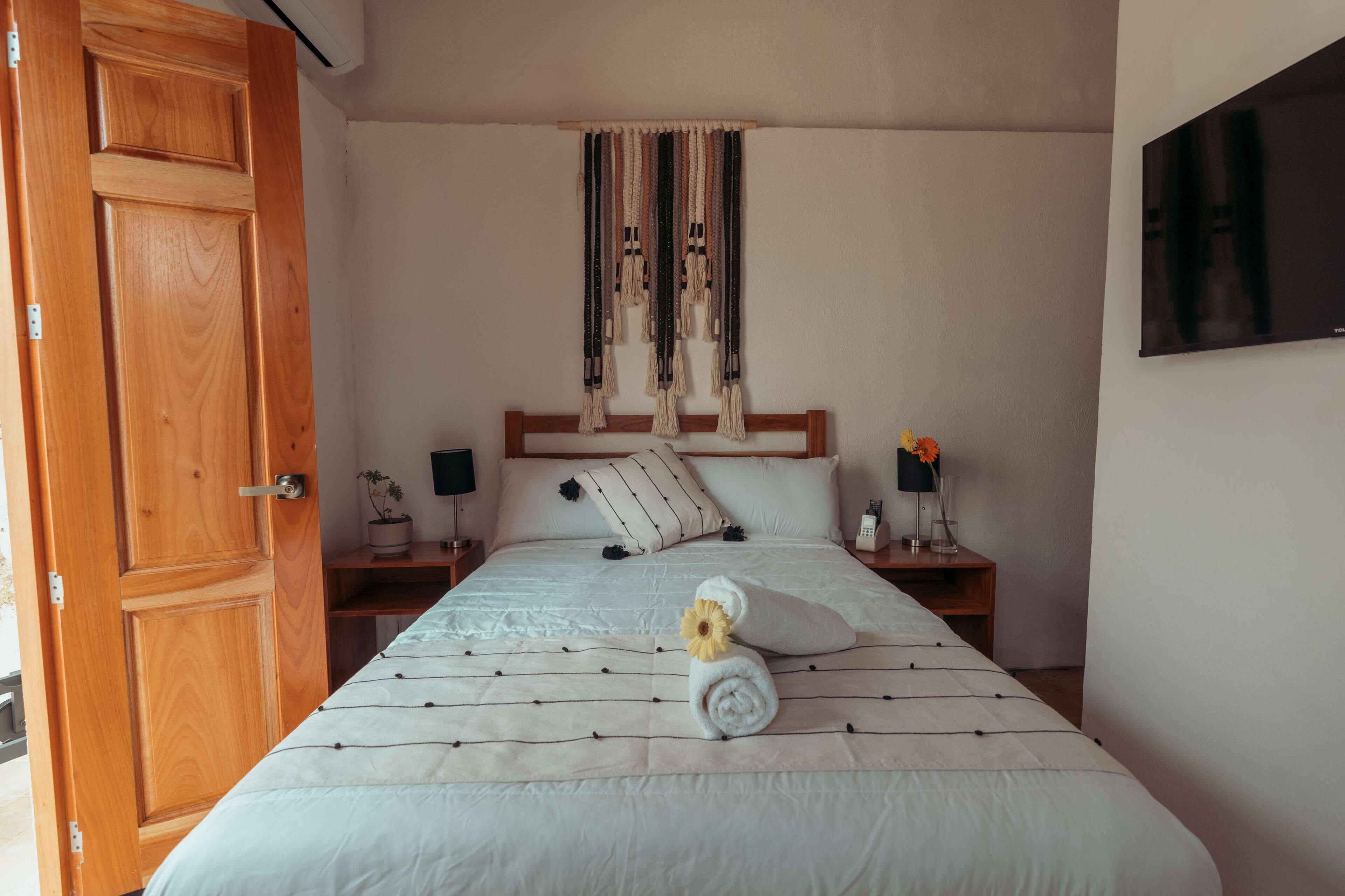 Ein ordentlich bezogenes Bett mit einem zusammengerollten Handtuch und einer Blume, eine Holztür, ein Nachttisch mit einer Lampe und ein an der Wand montierter Fernseher.