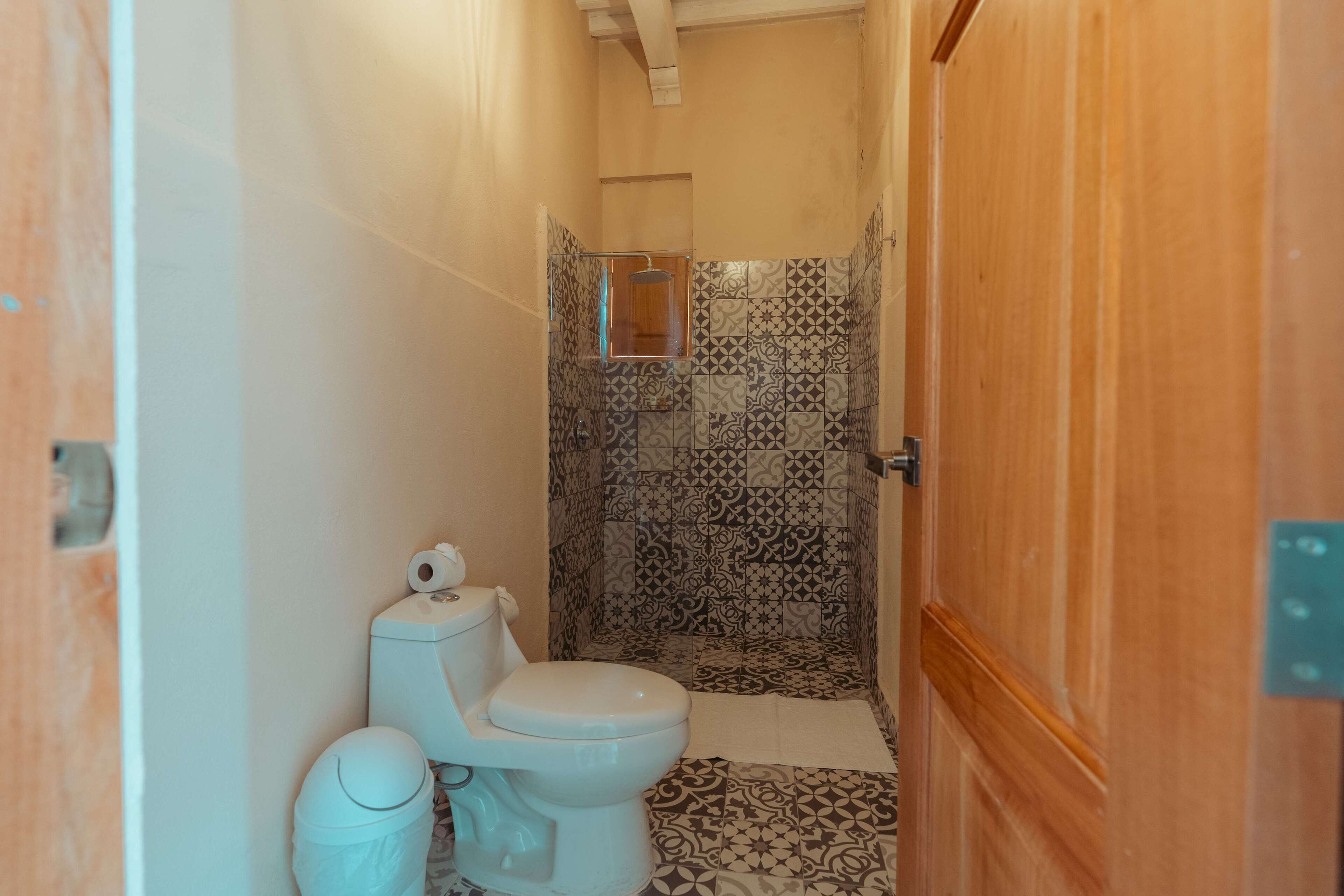 A small bathroom with a toilet, a trash can, and a shower area with patterned tiles.