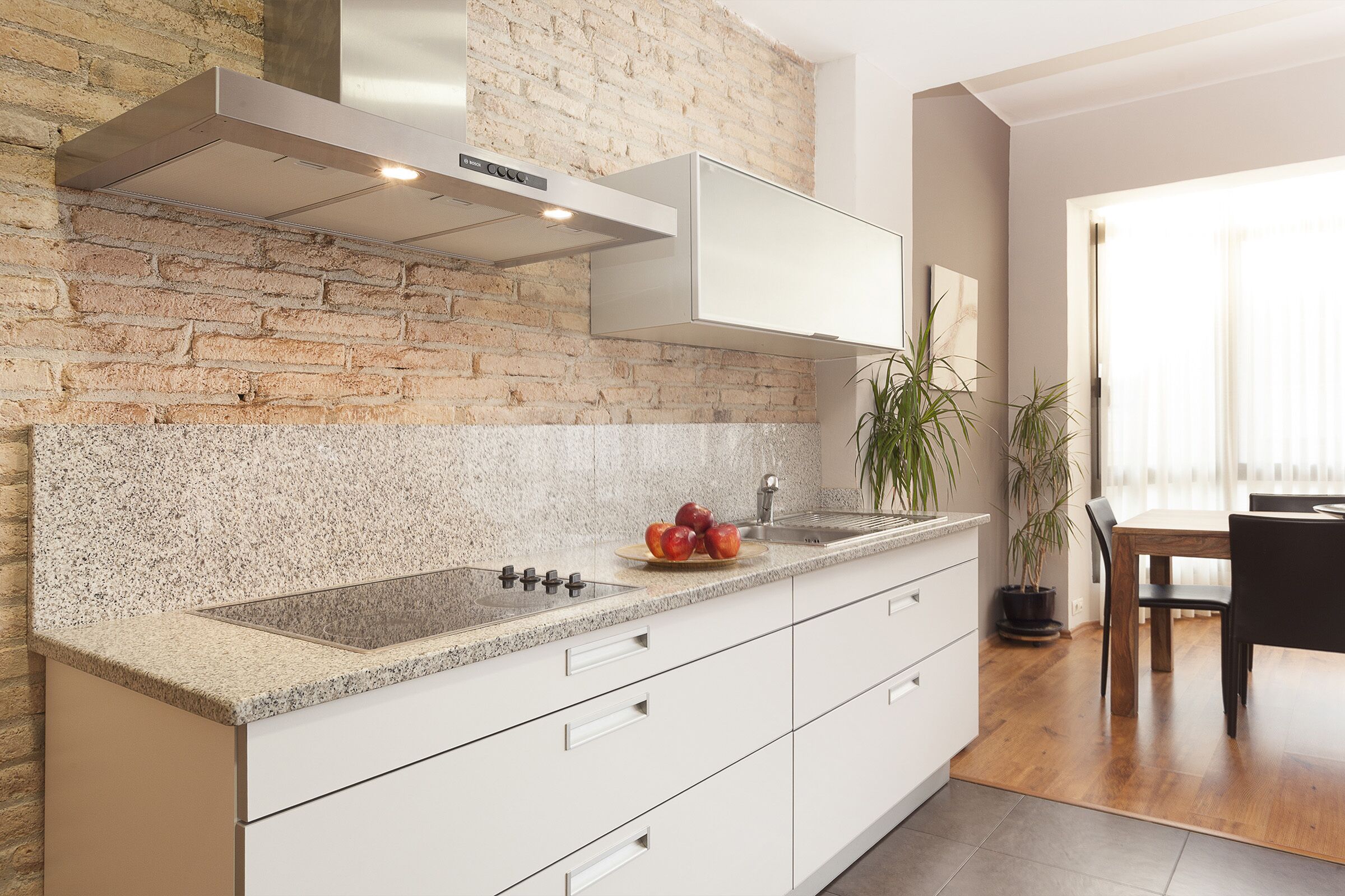 Una cocina moderna con encimera de granito, armarios blancos, una pared de acento de ladrillo y una campana extractora de acero inoxidable.