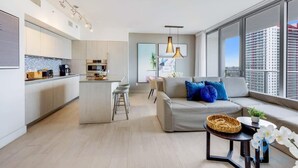 Un appartamento moderno con cucina, zona pranzo e soggiorno. Dispone di un'ampia finestra con vista sulla città, un divano con cuscini e un piccolo tavolino.