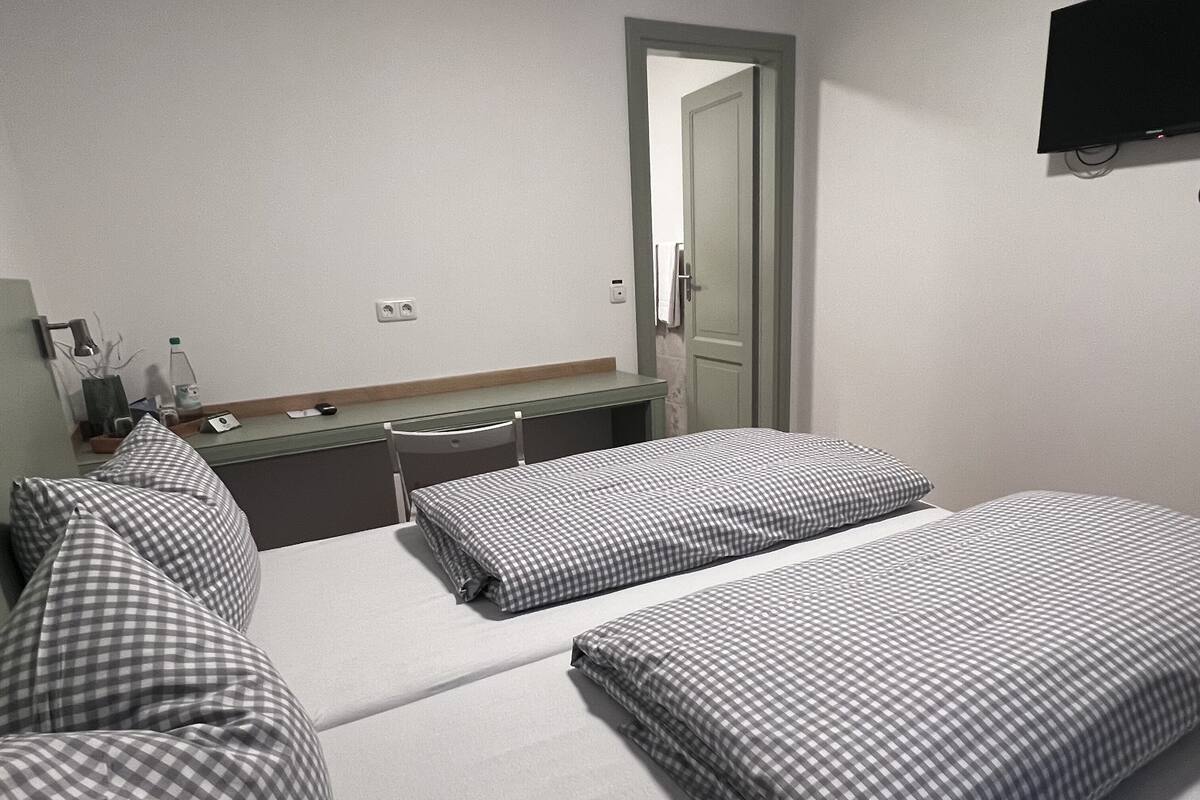 Ein Hotelzimmer mit zwei Betten, einem Schreibtisch, einem Stuhl, einem flachbildfernseher an der Wand und einer Tür zu einem anderen Raum.