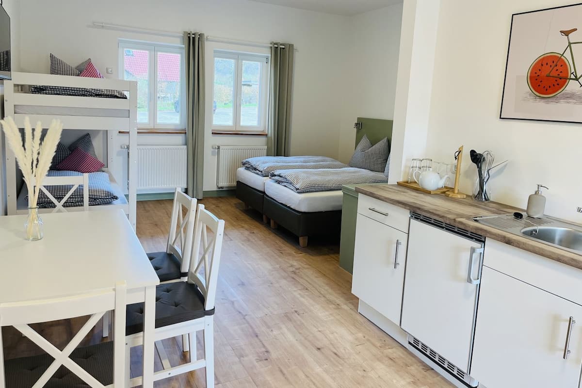 Ein kleines Zimmer mit Etagenbett, einem Einzelbett, einem Esstisch mit Stühlen, einer Küchenzeile mit Spüle und Geschirr sowie einem Fenster mit Vorhängen.