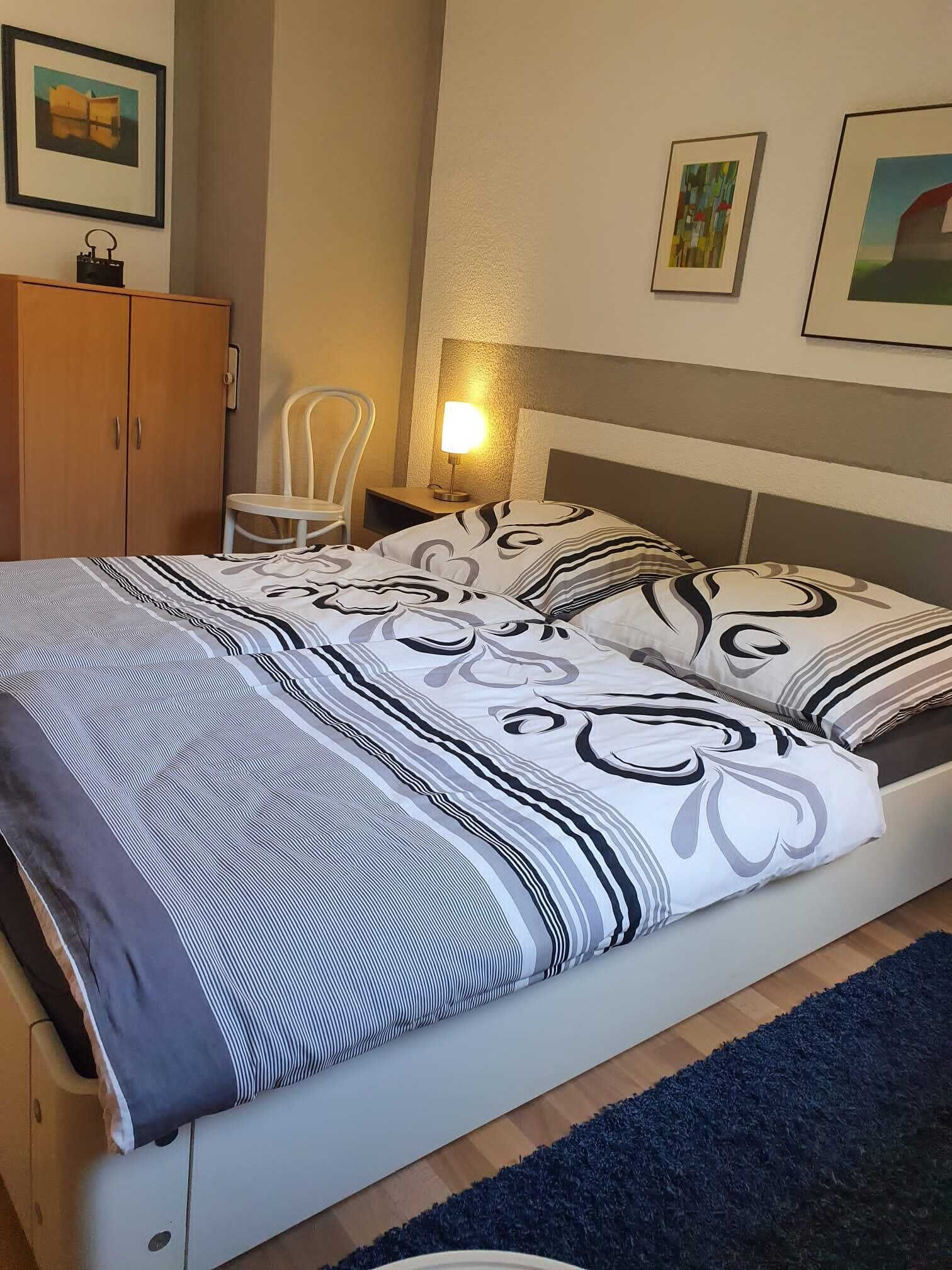 A neatly made bed with patterned bedding, a bedside lamp, a chair, and a wardrobe in a room with a simple, clean design.