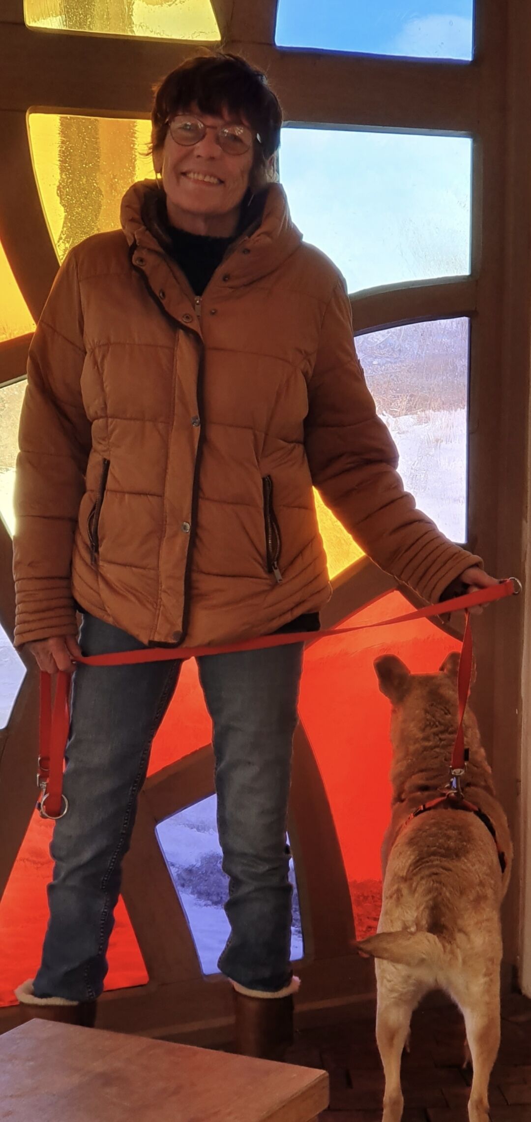 A person standing indoors with a dog on a leash, wearing a brown jacket and jeans.