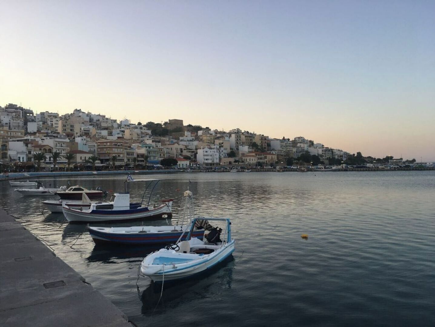 Una marina con diverse imbarcazioni ormeggiate, un fronte mare tranquillo e un contesto urbano fitto di edifici.