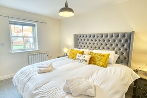 Cama bem arrumada com lençóis brancos e um travesseiro "Let's Nuggle", cabeceira cinza com capitonê, luminária de cabeceira e janela com persianas.