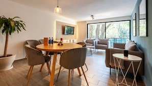 Ein modernes Wohnzimmer mit einem runden Holzesstisch, grau gepolsterten Stühlen, einer Topfpflanze und einem großen Fenster mit Blick auf Bäume.