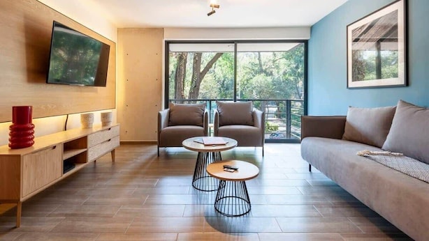 Ein modernes Wohnzimmer mit einer Couch, zwei Sesseln, einem Couchtisch, einem an der Wand befestigten Fernseher und einer großen Schiebeglasdoor zu einem Balkon.