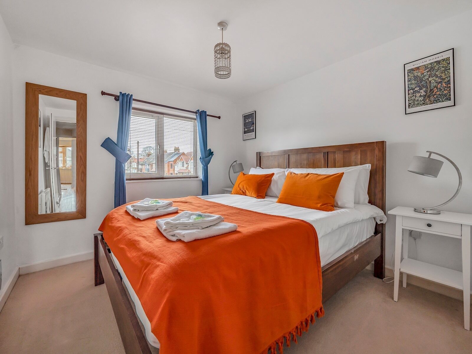 A neatly arranged bedroom with a bed, a bedside table, a nightstand, a window with curtains, a hanging light fixture, and a framed picture on the wall.