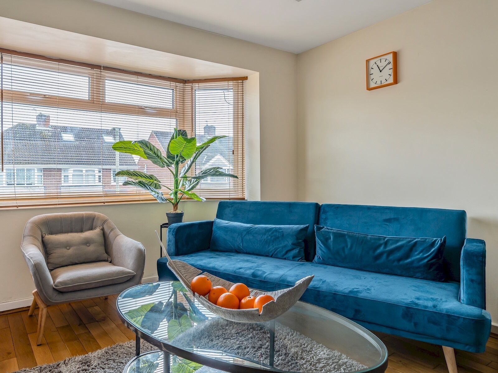 A living room with a blue sofa, a grey armchair, a glass coffee table with oranges, and a potted plant.