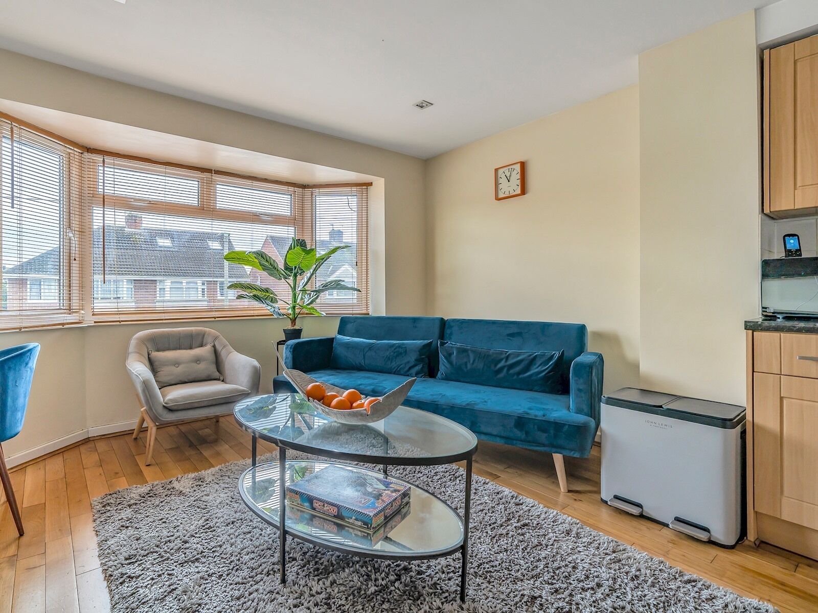 A modern living room with a blue sofa, a glass coffee table, a grey armchair, a potted plant, and a small kitchen area with a refrigerator.