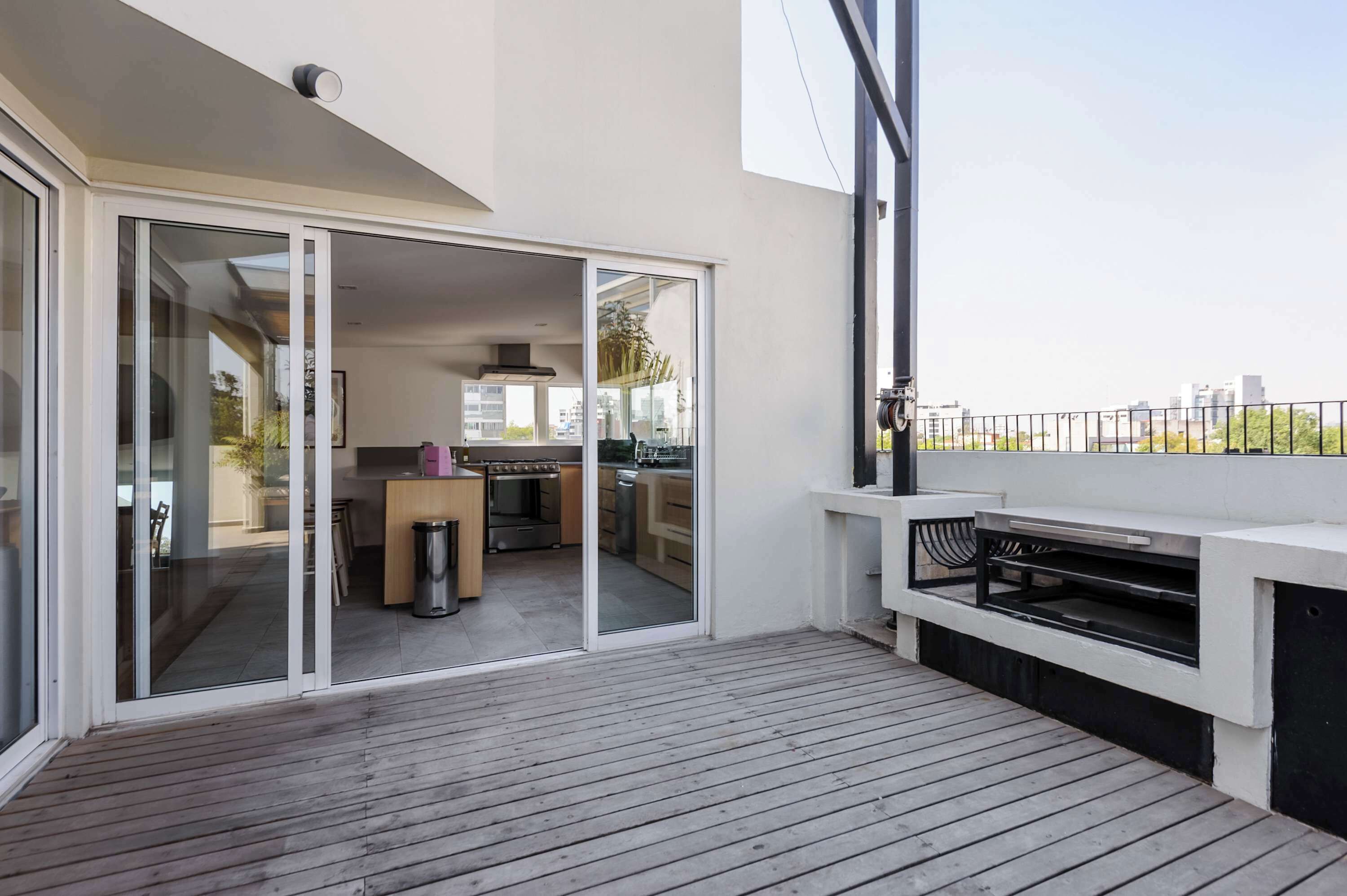 Un balcón moderno con piso de madera, una parrilla empotrada y una puerta corrediza de vidrio que da a una cocina interior.
