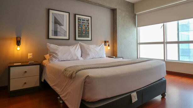 A modern hotel room with a large bed, two bedside tables, a nightstand, and a large window providing natural light. - w*| Lush 2BR w/AC Near Kennedy Park in Miraflores (Miraflores)