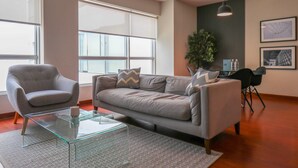 A modern living room with a grey sofa, a glass coffee table, and a grey armchair. There are large windows providing natural light and a plant in the corner. - w*| Lush 2BR w/AC Near Kennedy Park in Miraflores (Miraflores)