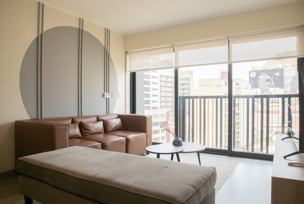 A modern living room with a sofa, a small round table, and a large window with a view of buildings.