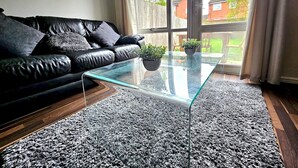 En moderne stue med et glas sofabord, en sort lædersofa og en grå, lurvet måtte.