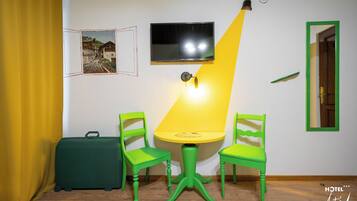 A room with a green suitcase, two green chairs, a yellow table, a TV mounted on the wall, and a framed picture.