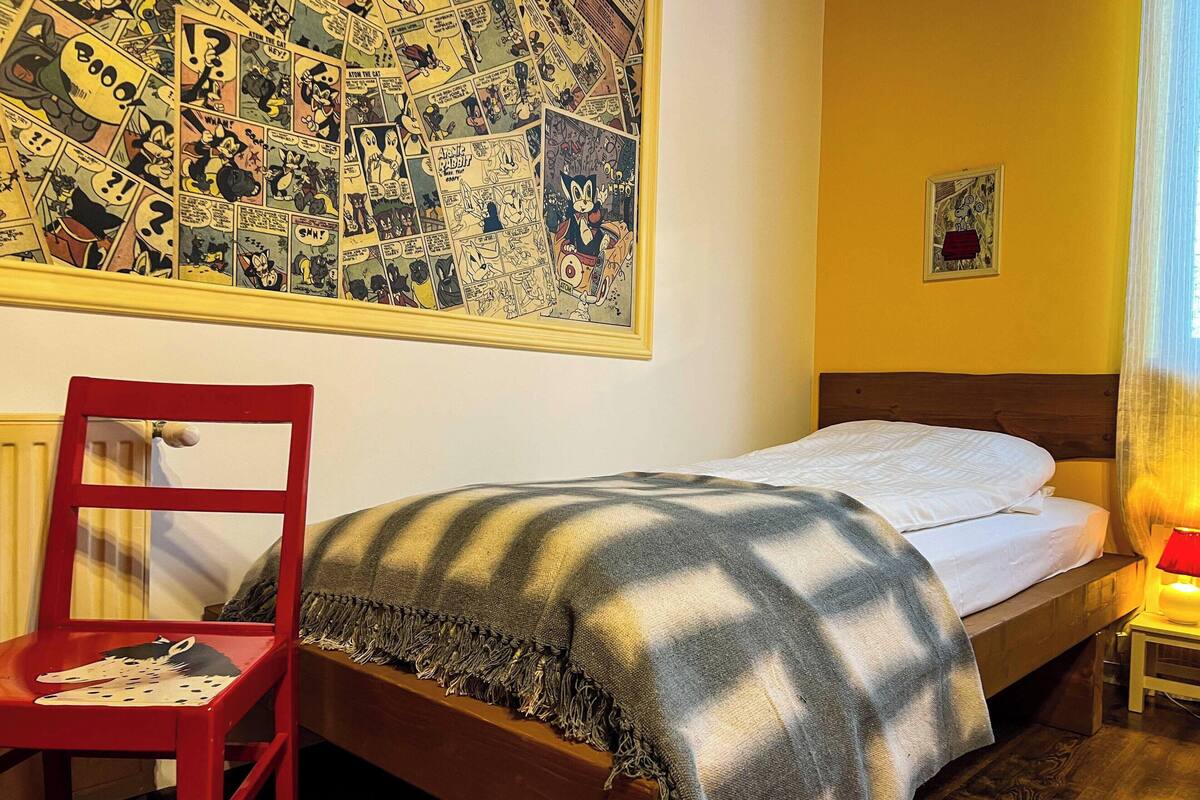 A room with a bed, a red chair, a wall with comic strip art, and a small table with a lamp.