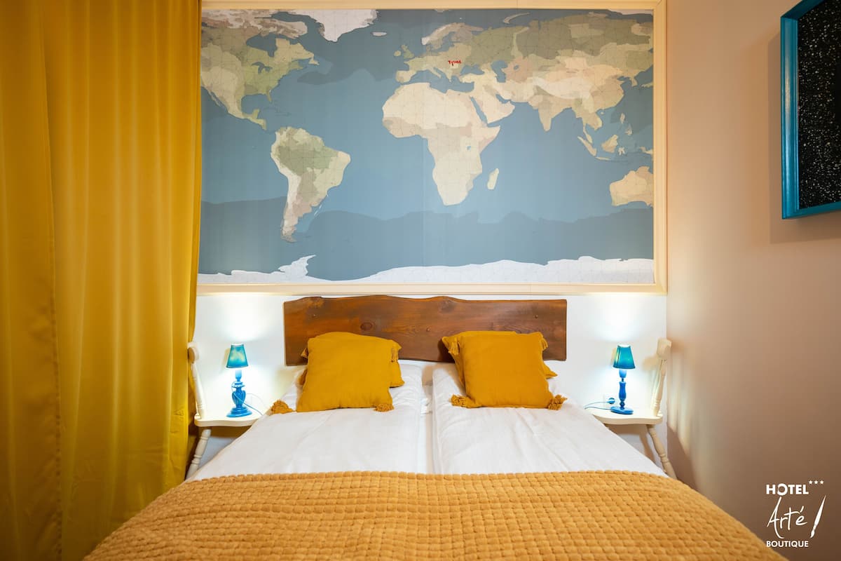 A hotel room with a large world map on the wall, a wooden headboard, two bedside tables with lamps, and a bed with a yellow blanket.