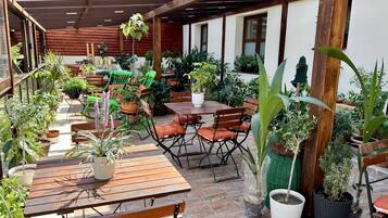 Un patio al aire libre con mesas y sillas de madera, rodeado de plantas en macetas y una pérgola de madera.
