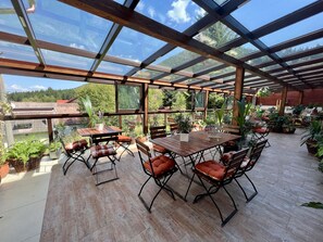 An outdoor patio with wooden tables and chairs, surrounded by potted plants and a glass ceiling.