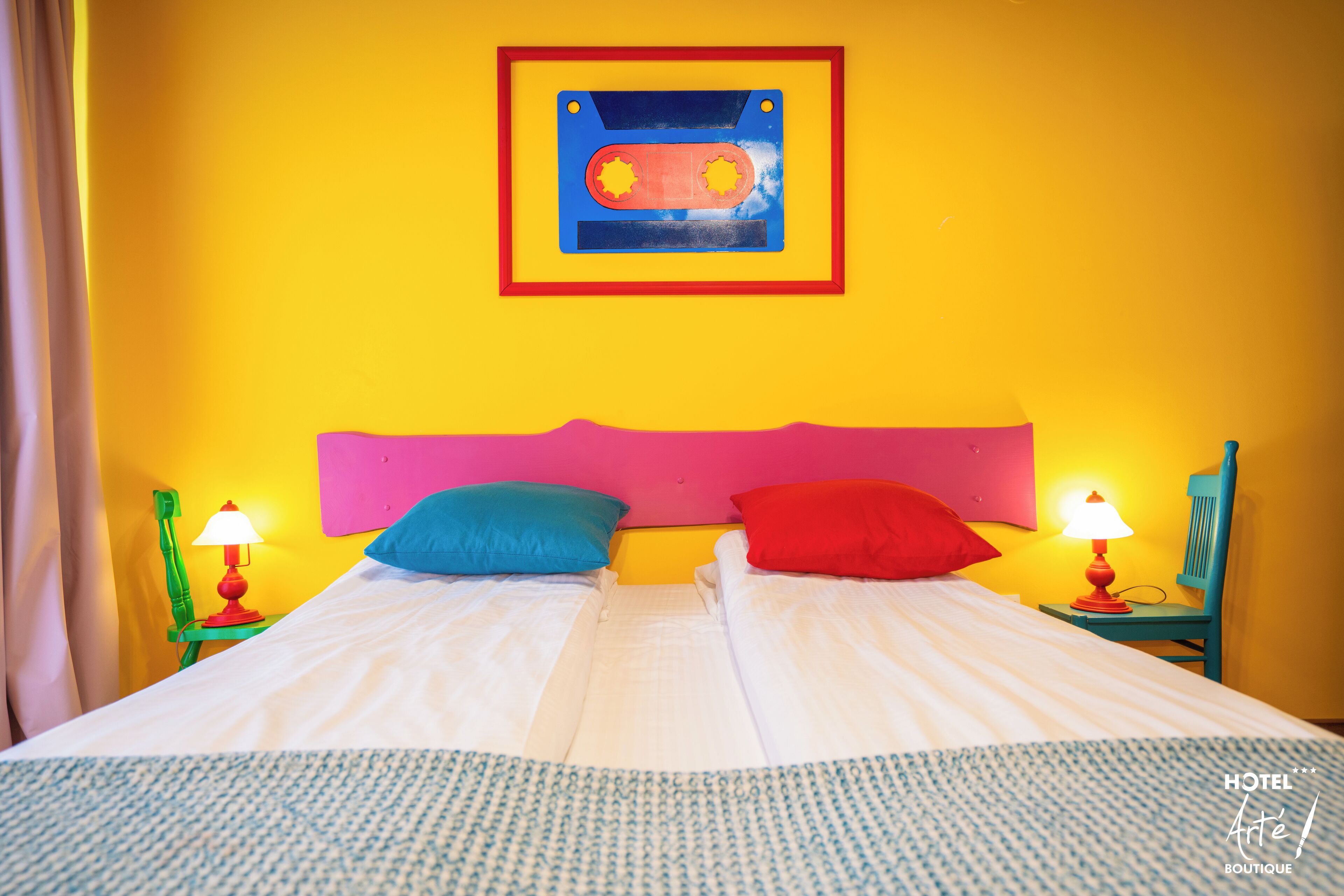 Un lit double avec du linge de lit blanc, deux oreillers, une œuvre d’art encadrée au-dessus, et deux tables de chevet avec des lampes.