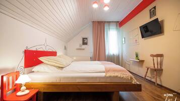 A bedroom with a wooden bed, a red chair, a bedside table with a lamp, a television on the wall, and a small shelf with a plant.
