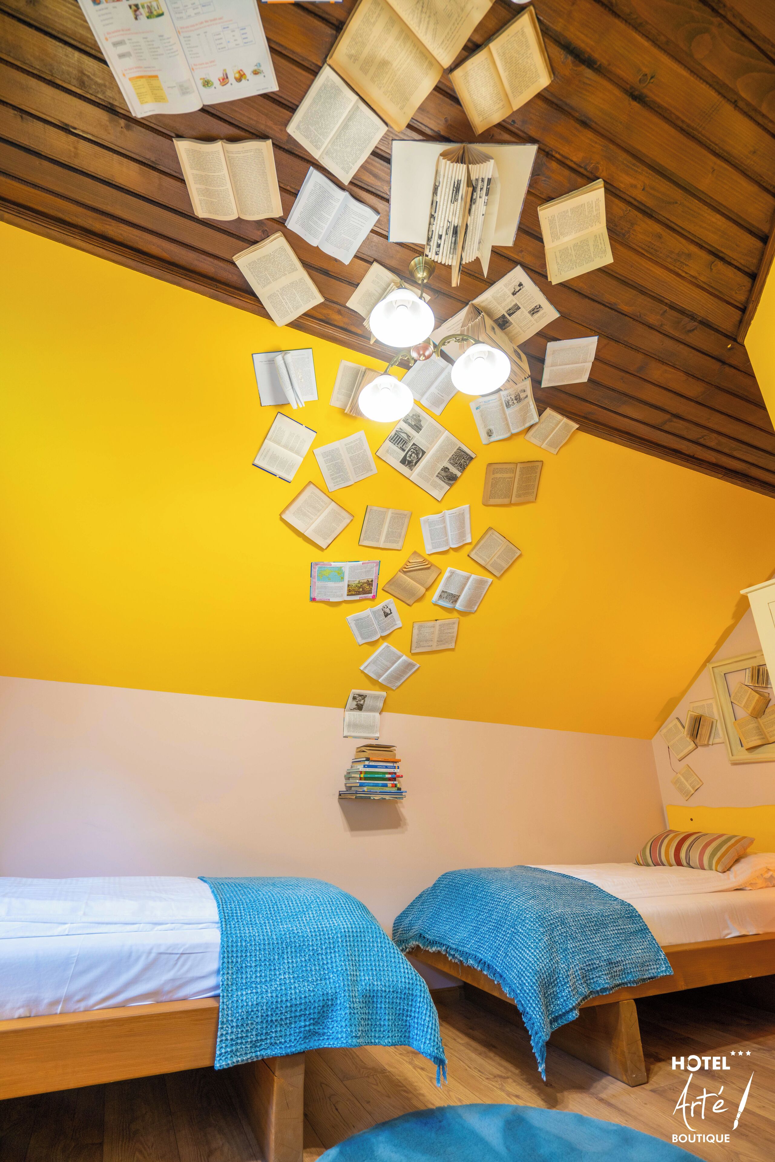 Una habitación con dos camas, suelo de madera y techo decorado con papeles y libros dispuestos en un diseño.