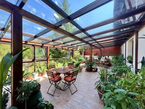 Ein überdachter Patio-Bereich mit Holztisch und -stühlen, umgeben von Blumentöpfen und Blick ins Grüne.