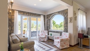 A cozy living room with a sofa, two floral armchairs, a small table, and a view of the outdoors through large windows. - Villa St. Nicholas House, Family Friendly, Large Heated Pool, 300m to the beach. (Corfu)