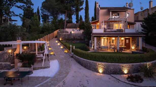 A two-story house with a stone facade, a balcony, and a well-lit outdoor area with seating and a ping pong table. - Villa St. Nicholas House, Family Friendly, Large Heated Pool, 300m to the beach. (Corfu)