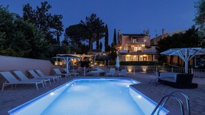 A swimming pool with lounge chairs and umbrellas, surrounded by a landscaped garden and a building with multiple balconies. - Villa St. Nicholas House, Family Friendly, Large Heated Pool, 300m to the beach. (Corfu)