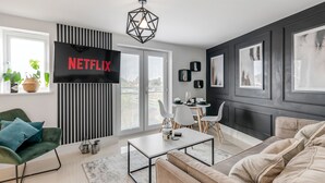 A modern living room with a large flat-screen TV displaying 'NETFLIX', a green armchair, a beige sofa, a white coffee table, and a geometric pendant light. - Modern Apartment (Wymondham)
