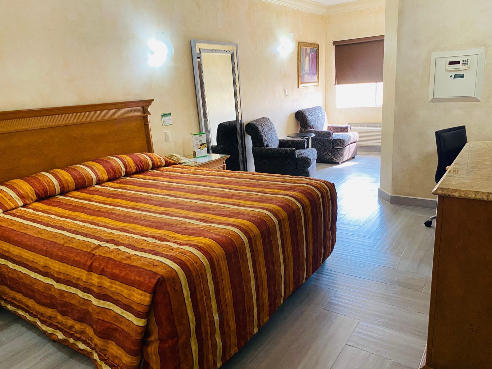 Una habitación de hotel con una cama grande, un escritorio, dos sillones, un espejo y un termostato en la pared.