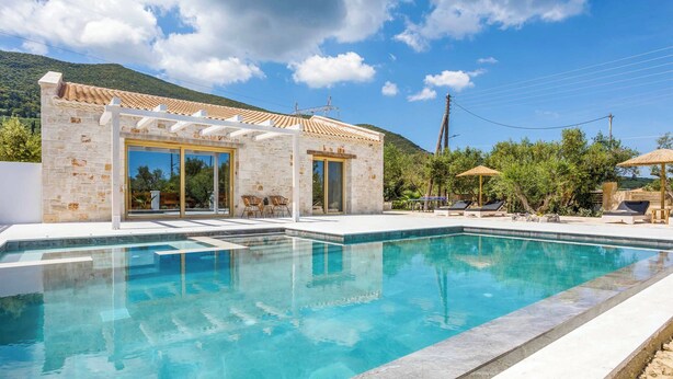 A modern pool area with a stone building, outdoor seating, and a mountainous backdrop.