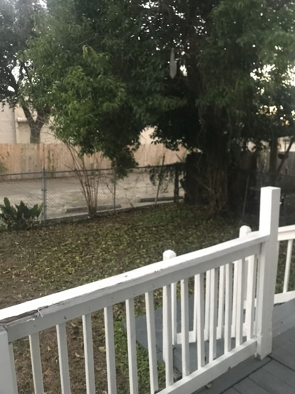 Uma cerca de metal branca com barras verticais, um espaço pavimentado com alguma vegetação e um edifício ao fundo.