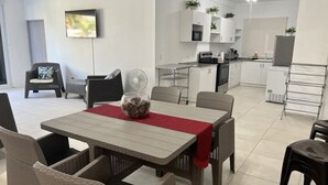Une cuisine moderne avec des meubles blancs, un four à micro-ondes et une machine à café. On y trouve un coin repas avec des chaises en osier et une table recouverte d’une nappe rouge.