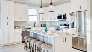 A modern kitchen with white cabinets, stainless steel appliances, and a central island with bar stools. - Luxurious 7BR Beach Home w/ Ocean Views, Pool, Hot tub, and Elevator (North Topsail Beach)