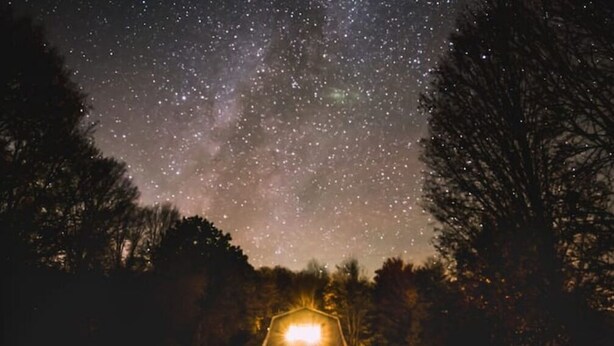 Eine ländliche Nachtszene mit einem beleuchteten Bauernhof, umgeben von Bäumen und einem Sternenhimmel.