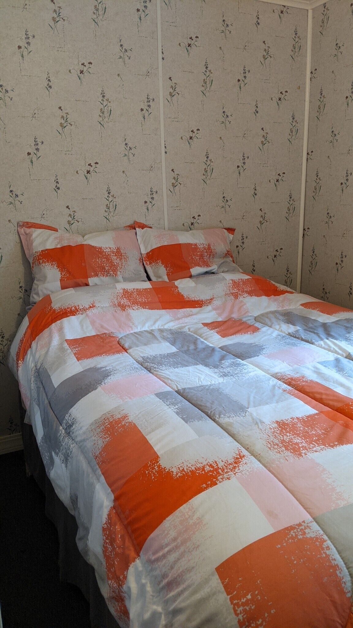 En säng med ett quiltat täcke i orange, vitt och grått, och en blommig tapet på väggarna.