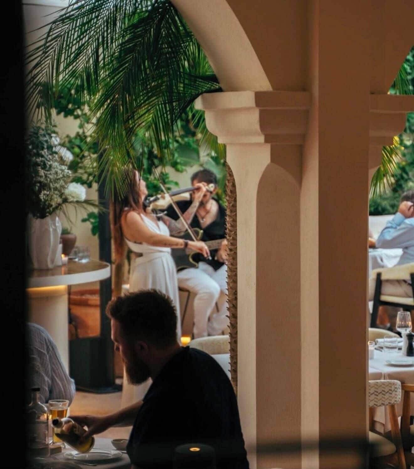 An outdoor dining area with a covered section, palm trees, and a person playing a guitar.